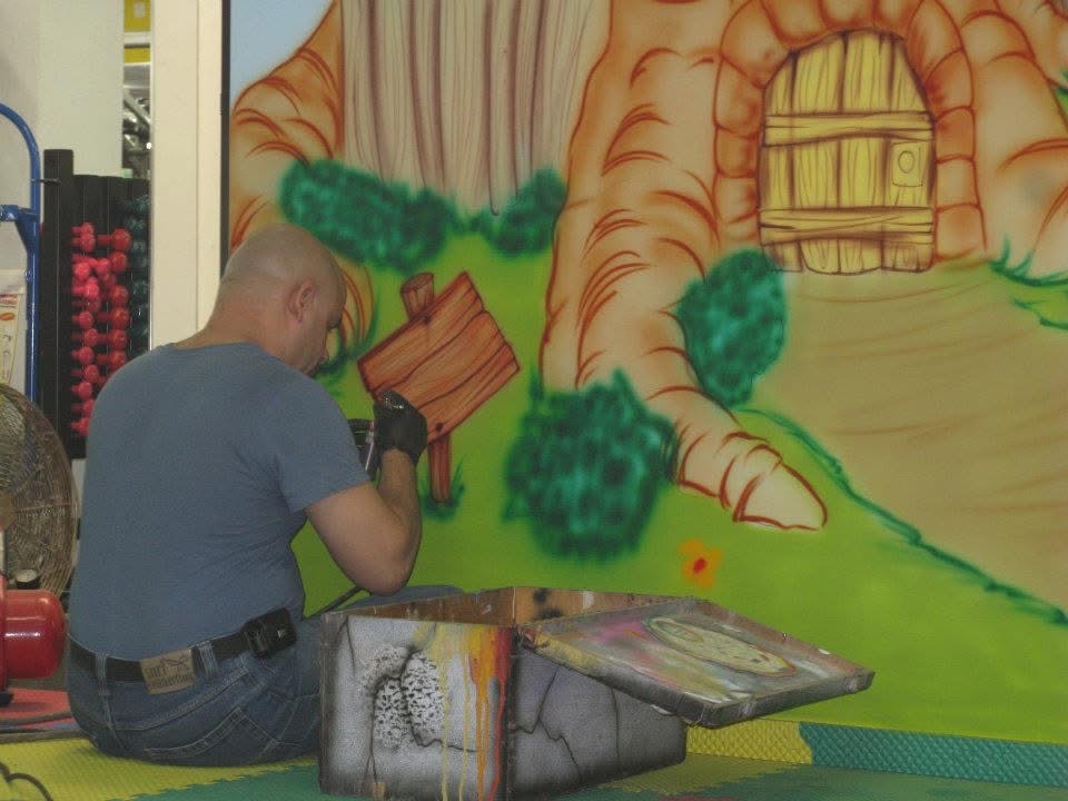 Efrain airbrushing a storybook tree house scene freehand onto a wall
